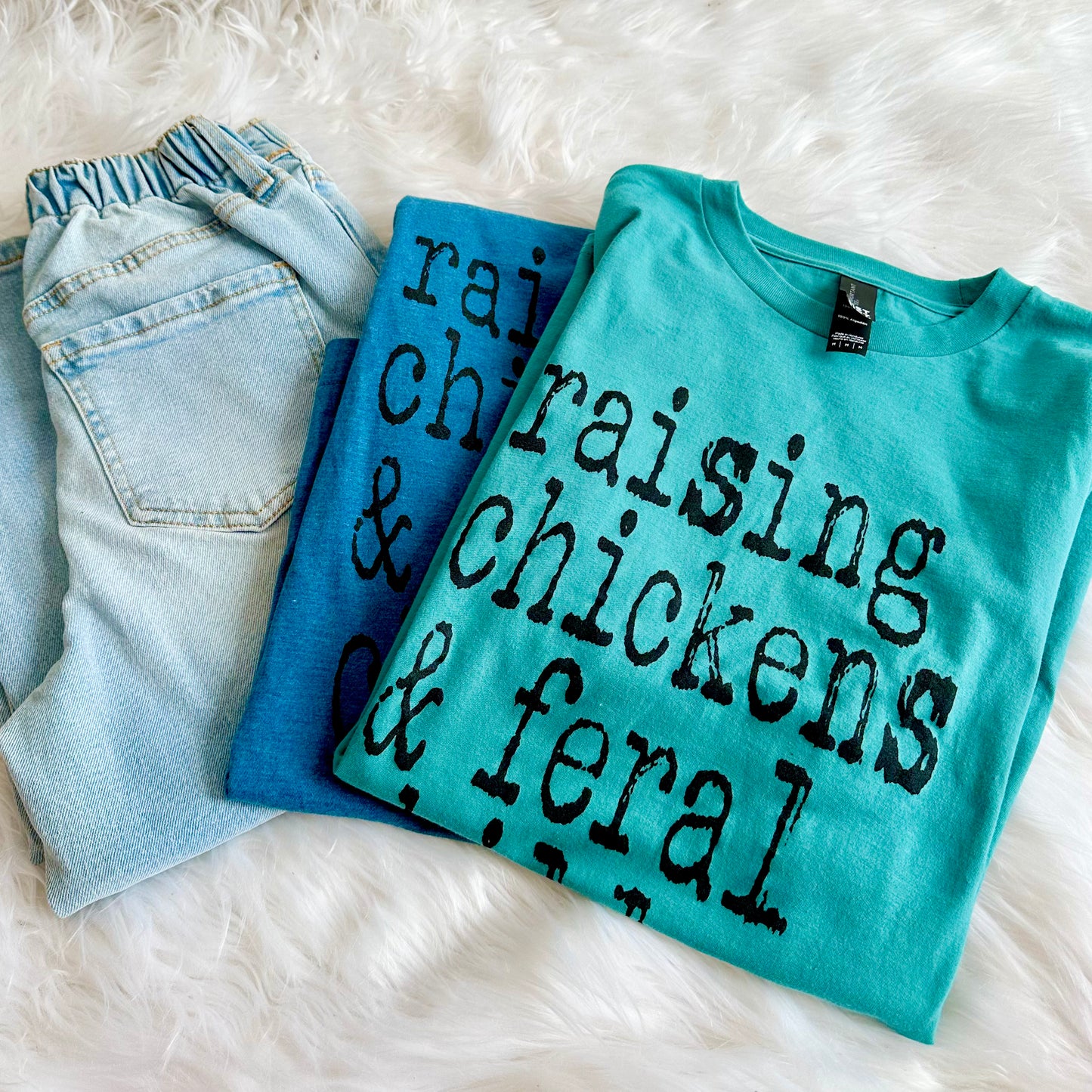 Folded blue jeans and two turquoise t-shirts with text on a white fluffy surface