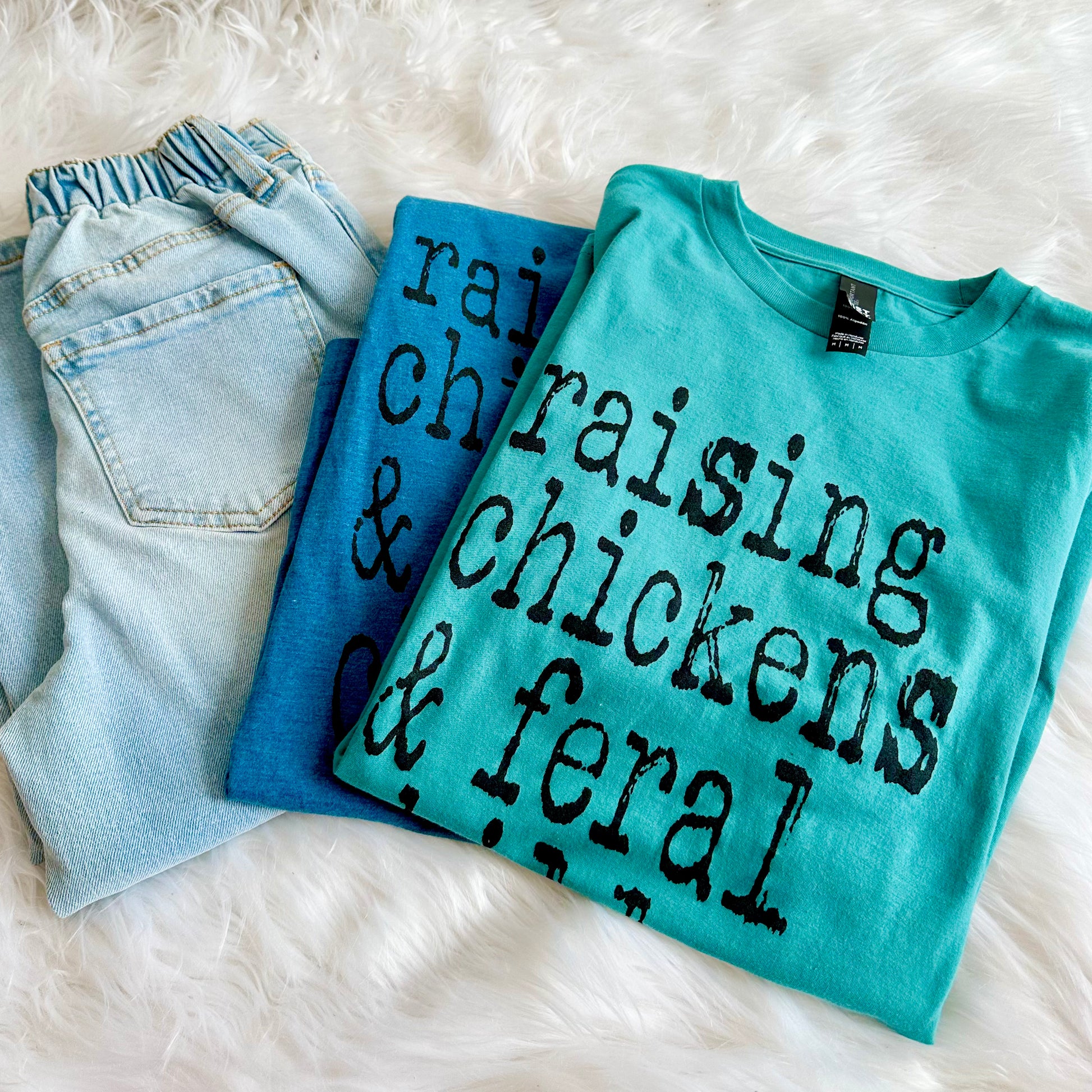 Folded blue jeans and two turquoise t-shirts with text on a white fluffy surface