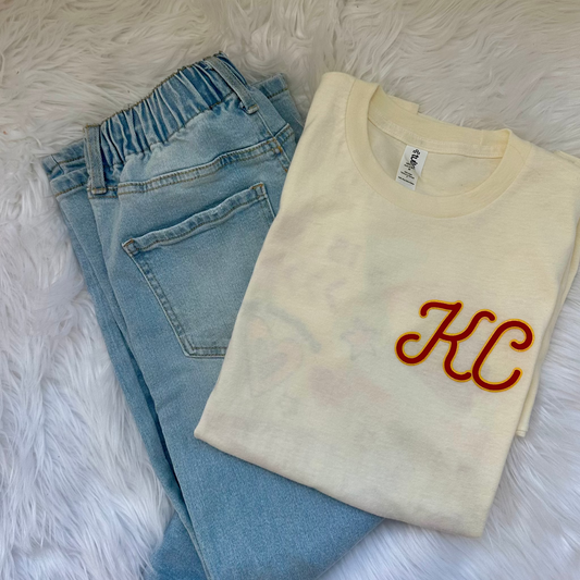 Folded cream t-shirt with red and yellow "KC" design next to light wash jeans on a fluffy white background. 