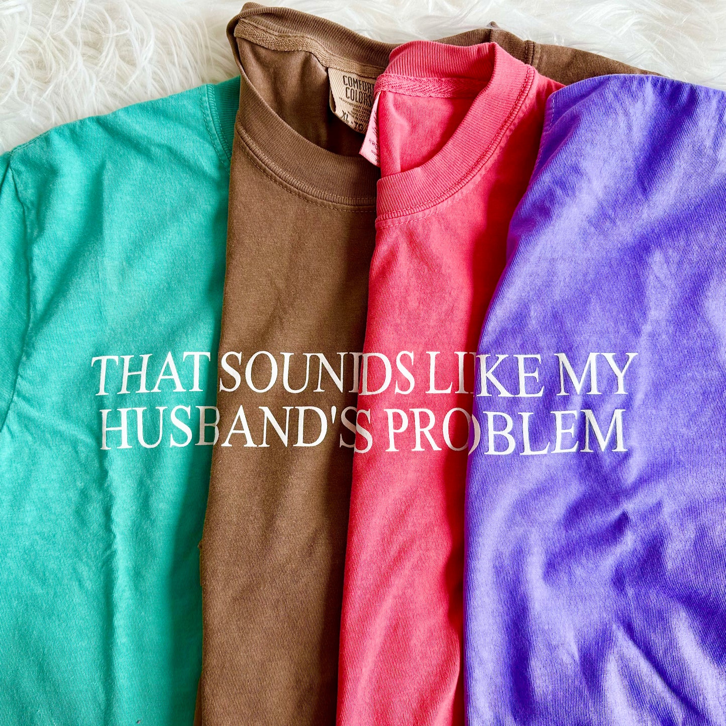 Four folded t-shirts in teal, brown, red, and purple with text 'THAT SOUNDS LIKE MY HUSBAND'S PROBLEM' on a white background.