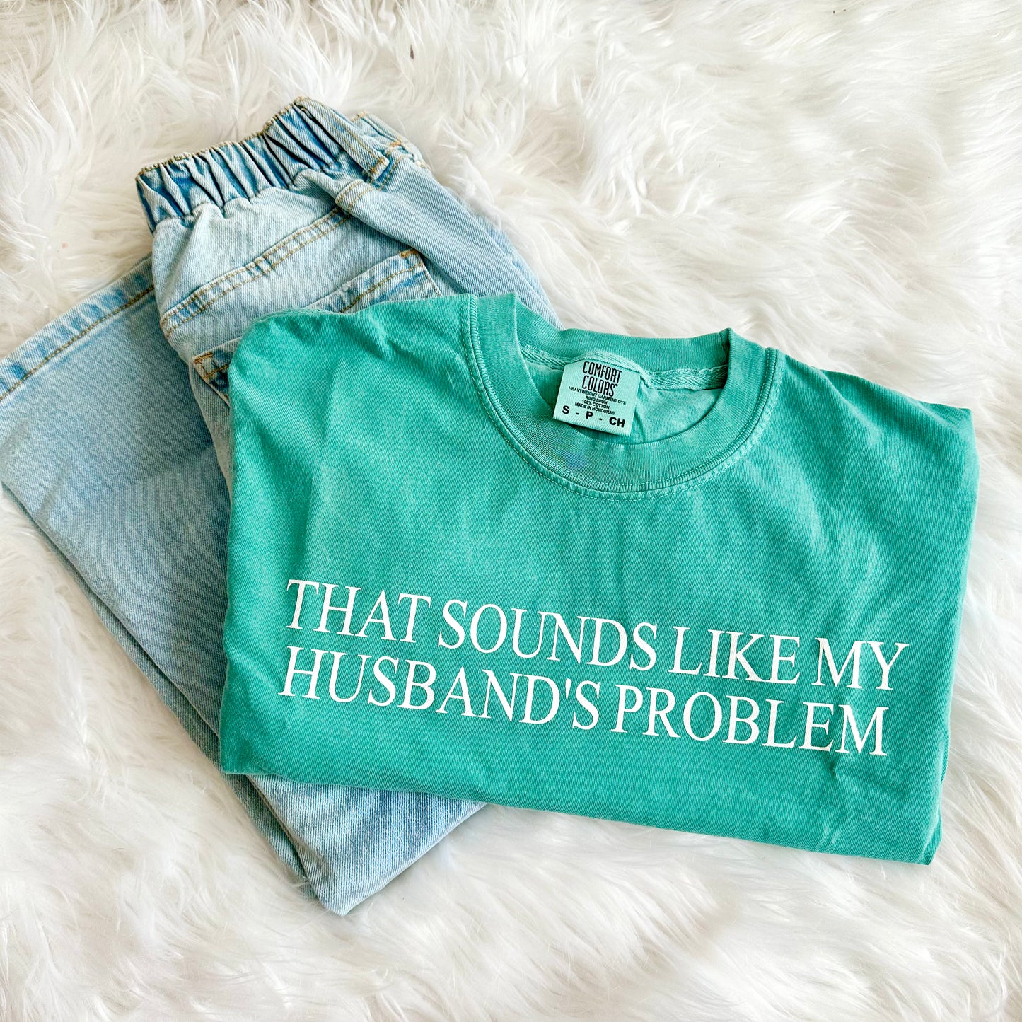 Teal t-shirt with text 'THAT SOUNDS LIKE MY HUSBAND'S PROBLEM' on a white fluffy surface