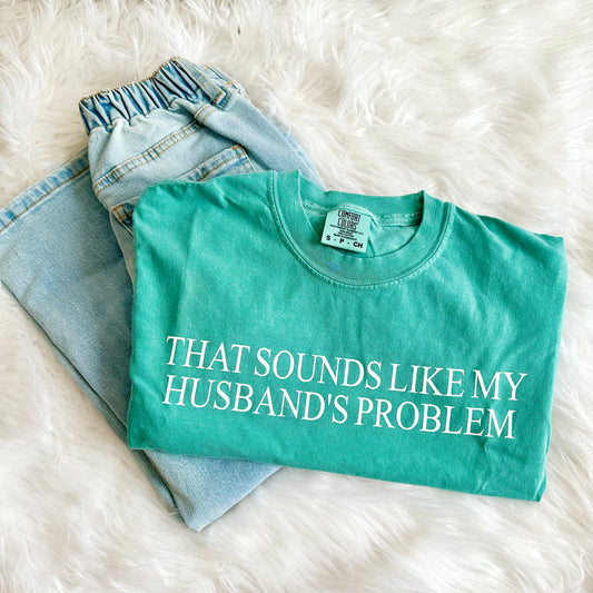 Teal t-shirt with text 'THAT SOUNDS LIKE MY HUSBAND'S PROBLEM' on a white fluffy surface