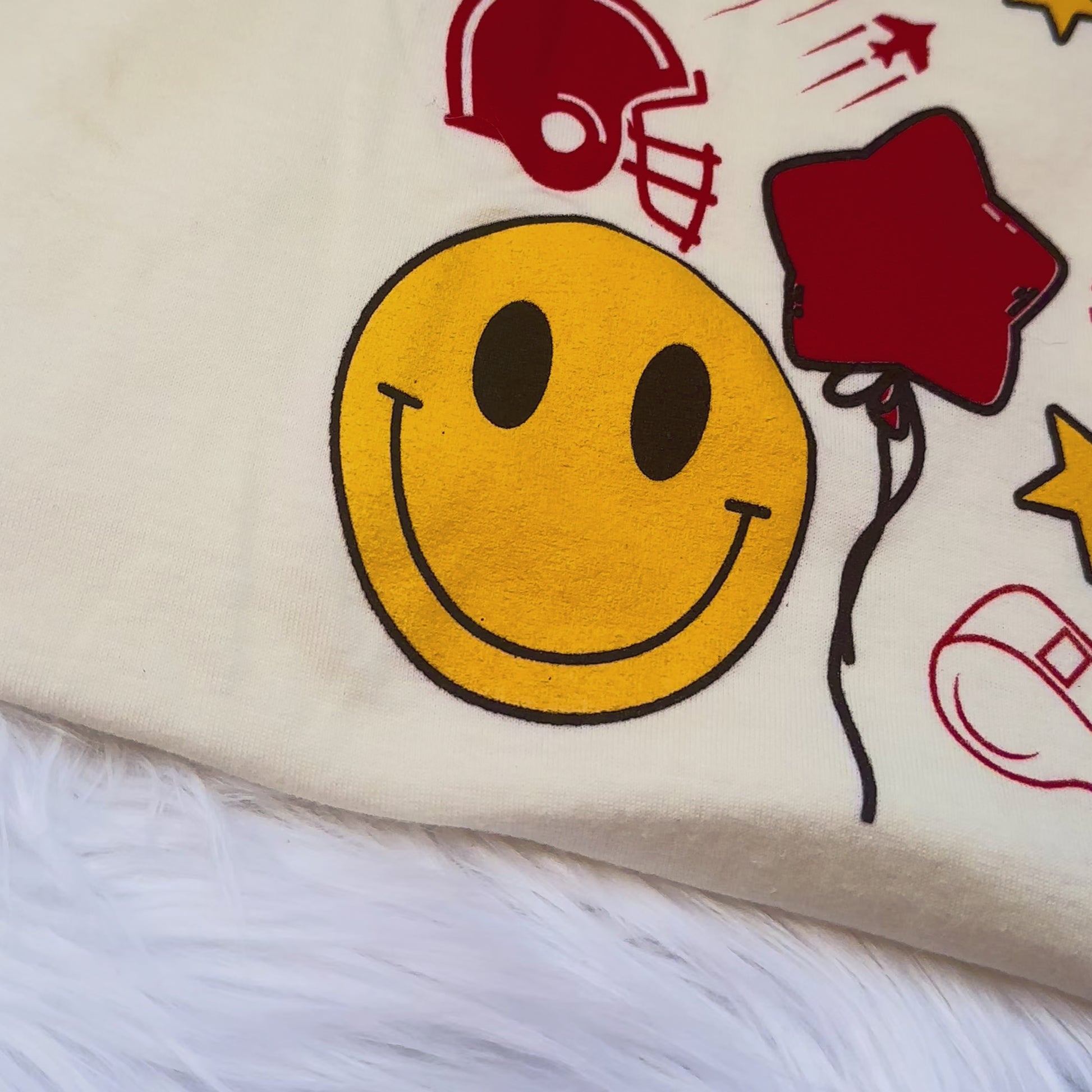 back of kansas city cream tshirt video with up close shots of the back collage design. The video shows a smiley face, balloon, football helmet, jets, whistle, banner, KANSAS CITY, hearts, stars, arrowhead, 15, 87, TOUCHDOWN, Establisjed 1980, lightning bolt, football, and play drawings. 