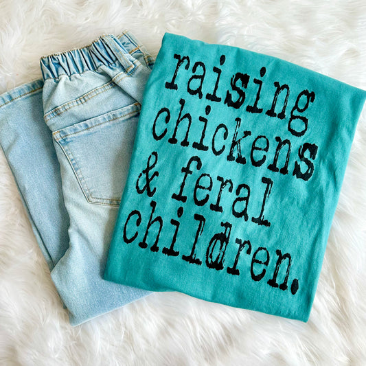 Teal shirt with text 'raising chickens & feral children' on a fluffy white surface
